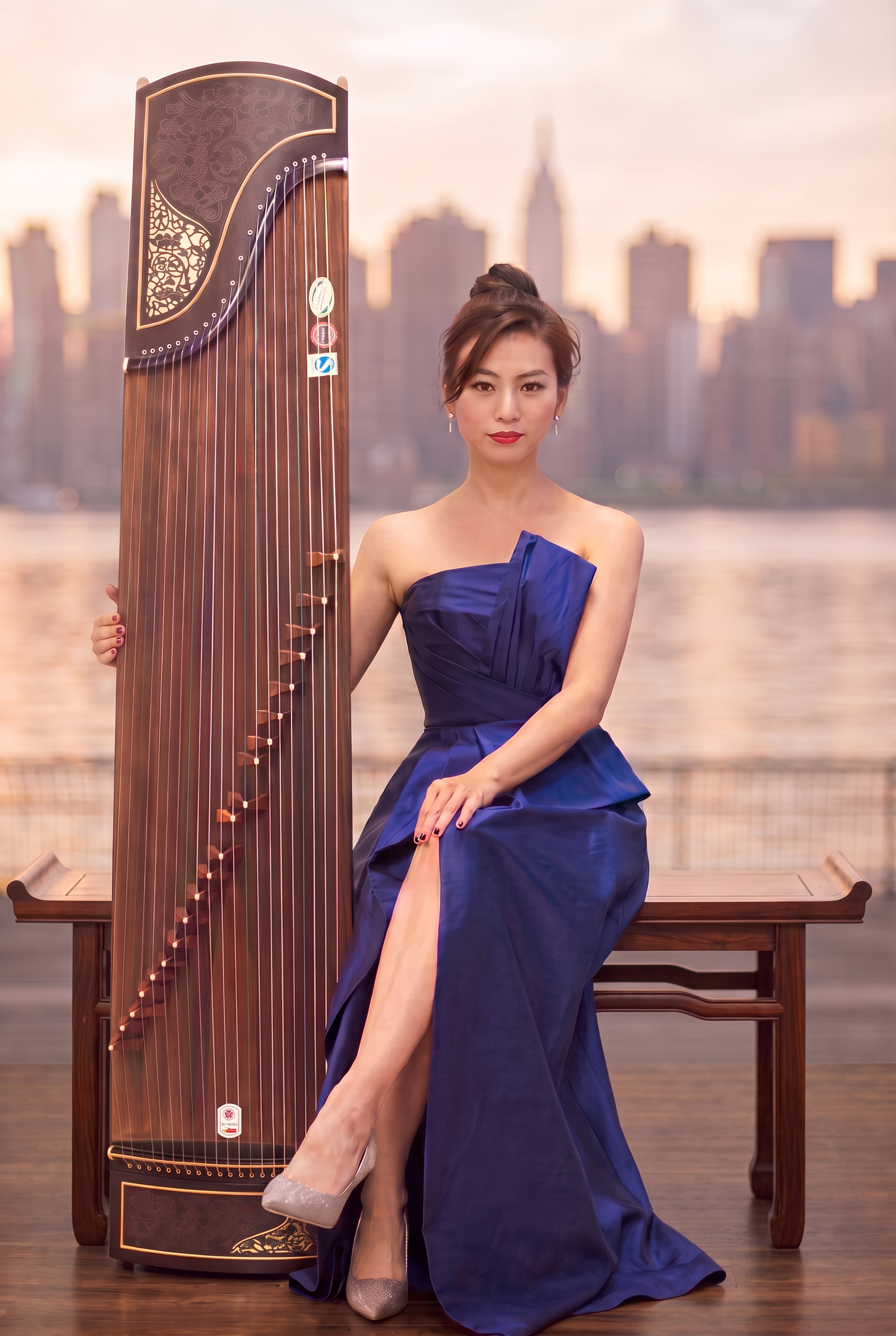 Wei Sun, New York guzheng performer, seated on a Ming-style wooden bench in a royal blue gown beside her guzheng, with a soft Midtown Manhattan skyline across the water